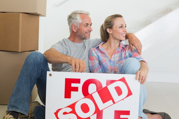 Feliz pareja sentada en el piso con cartel vendido