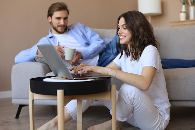 Feliz pareja sentada, relajándose en el piso de la sala de estar, usando la computadora portátil para comprar en línea juntos, viendo películas.