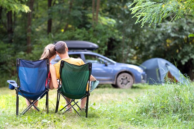 Feliz pareja sentada en sillas en el camping abrazándose junto con un coche y una tienda de campaña en el fondo.