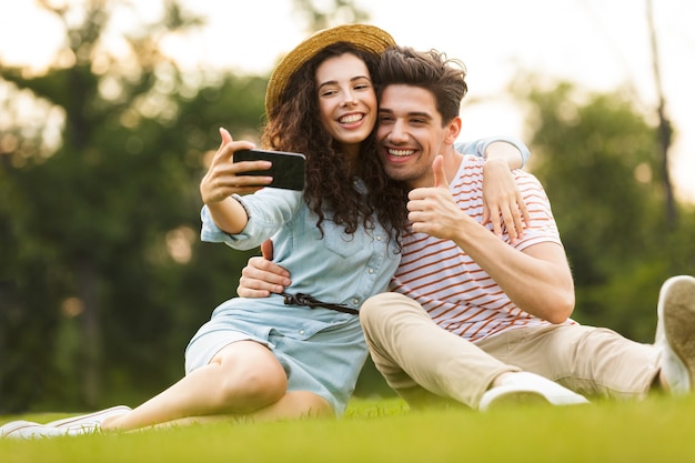 Feliz pareja sentada sobre la hierba verde en el parque y tomando selfie en smartphone