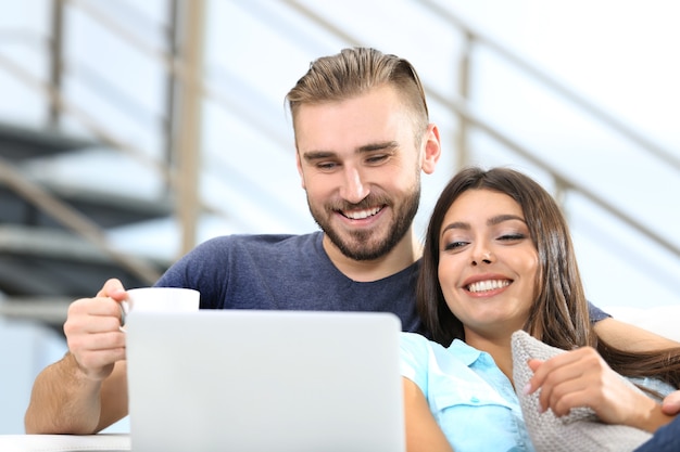 Feliz pareja sentada en el sofá con café y trabajando en una computadora portátil