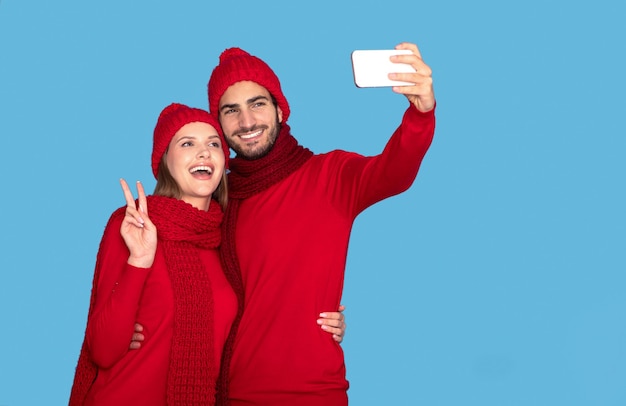 Feliz pareja con sombreros y bufandas de invierno tomando selfie en un teléfono inteligente