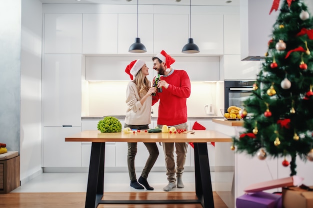 Feliz pareja con sombreros de santa en las cabezas brindando con cerveza mientras está de pie en la cocina en Nochebuena