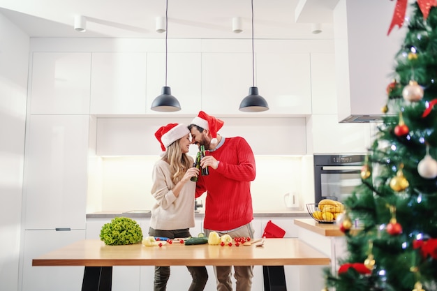 Feliz pareja con sombreros de santa en las cabezas brindando con cerveza mientras está de pie en la cocina en nochebuena