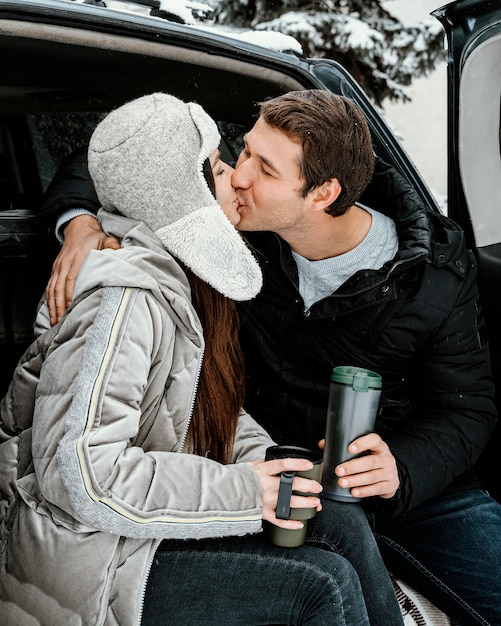 Feliz pareja tomando una bebida caliente en el maletero del coche y besándose durante un viaje por carretera