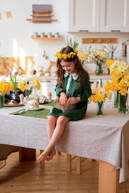 Feliz Páscoa alegre linda garota em um vestido verde com uma coroa de flores brinca com um coelho em casa na cozinha