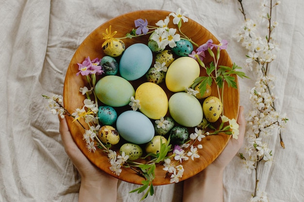 Feliz Páscoa Mãos segurando uma tigela de madeira com ovos de páscoa elegantes e flores de primavera florescendo na mesa rústica Páscoa rústica plana postura Ovos pintados naturais e flores em tecido de linho