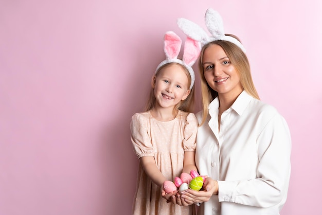 Feliz Páscoa Retrato de linda mãe loira e filha com orelhas de coelho e uma cesta de ovos coloridos em suas mãos Fundo rosa Espaço para texto Foto de alta qualidade