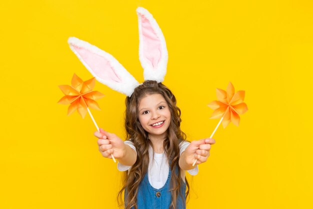 Feliz Páscoa Uma garotinha está comemorando alegremente um feriado de primavera Uma criança com orelhas na cabeça retrata um coelho Uma mocinha segura flores de origami em um fundo amarelo isolado