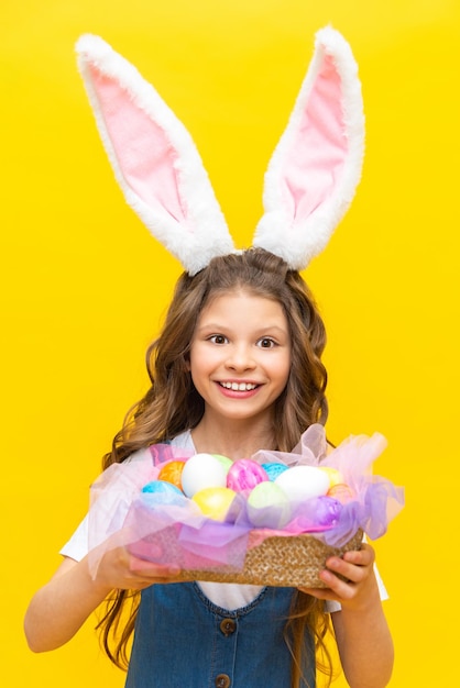 Feliz Páscoa Uma garotinha segura uma cesta de ovos coloridos para um feriado de primavera Uma criança encantadora com orelhas de coelho em um vestido de verão azul sobre um fundo amarelo isolado