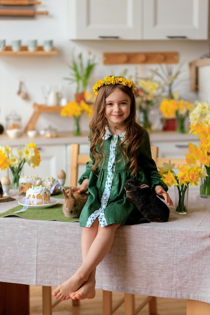 Feliz Pascua alegre hermosa chica con un vestido verde con una corona de flores juega con un conejo en casa en la cocina