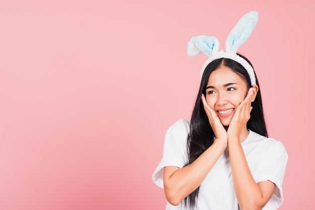 Feliz Pascua. Hermosa joven adolescente sonriendo con orejas de conejo de Pascua sosteniendo sus mejillas emocionada sorprendida, masaje facial femenino de retrato, toma de estudio aislada en fondo rosa