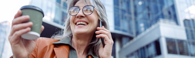 Feliz pensionista feminina com copo de bebida n fala no celular na rua da cidade moderna