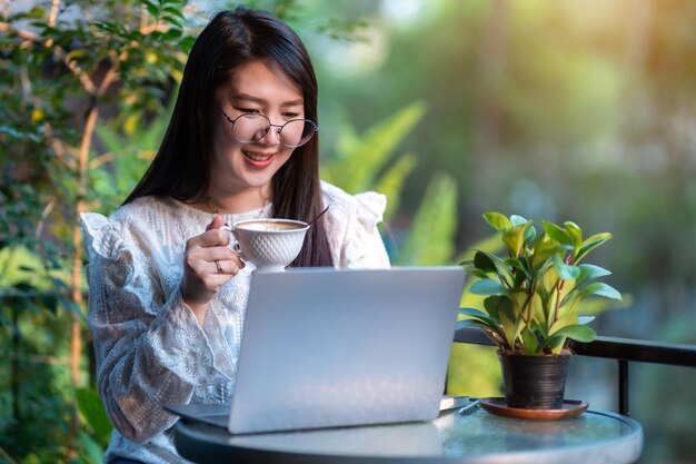 Feliz por mulheres de negócios freelance asiáticas, segure uma caneca de café casual trabalhando com laptop com Notebook e smartphone fora do café com árvores frondosas Estilo de vida empresarial