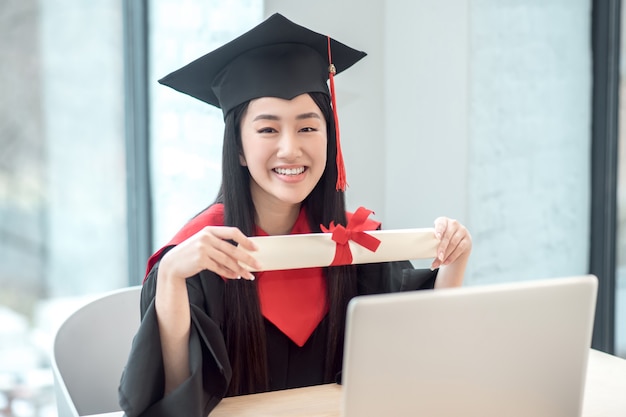 Feliz pós-graduação. fofa asiática sorridente graduada segurando o diploma e parecendo feliz