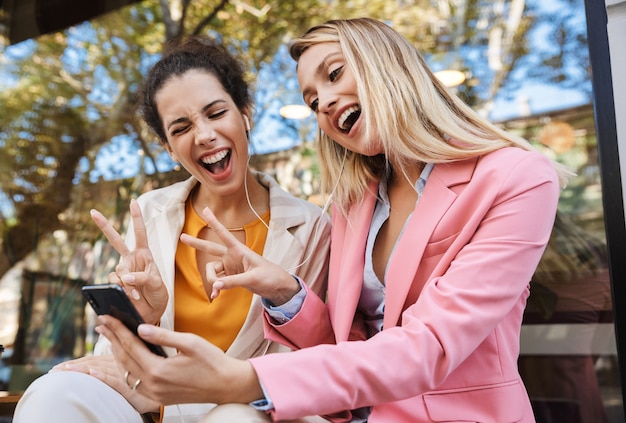 un feliz positivo dos colegas de mujeres de negocios al aire libre mediante teléfono móvil en auriculares.