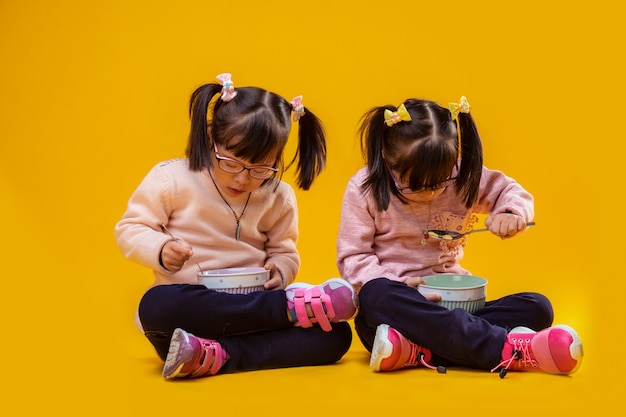 Feliz y positivo. Hermanitas bonitas atentas sentadas en el piso desnudo y comiendo cereales de cuencos hondos