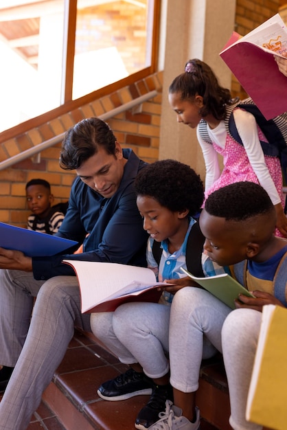 Foto feliz professor masculino diversificado e crianças com livros sentados nas escadas na sala de aula da escola primária