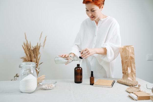 Feliz químico femenino pelirrojo preparando una mezcla de cosméticos ecológicos en una mesa. Agregar líquido en una botella de plástico marrón común con una pipeta