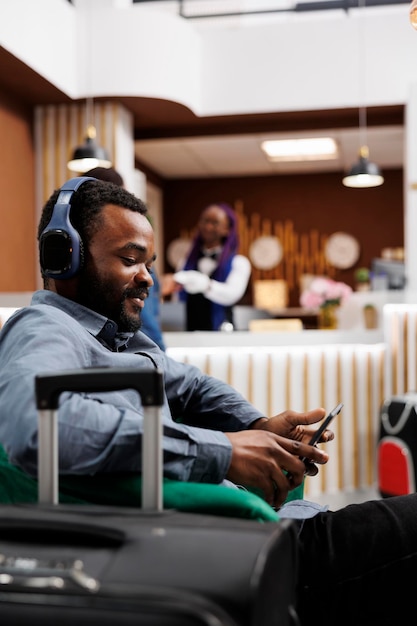 Feliz y relajado hombre afroamericano sentado en el vestíbulo del hotel escuchando música en una tableta digital, haciendo una videollamada mientras espera el check-in. Turista con auriculares mirando la pantalla del dispositivo