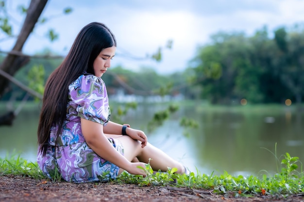 Feliz Relajante Retrato mujer asiática Use vestido púrpura con reloj inteligente mientras se sienta en el césped verde al lado de un embalse en el parque de la ciudad al aire libre