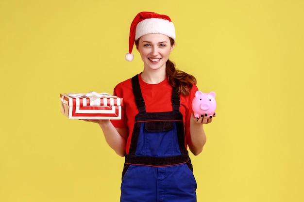 Feliz repartidora sosteniendo una caja de regalo envuelta y una alcancía, ahorros para el servicio de entrega en línea, vestida con un mono azul y un sombrero de santa claus. Disparo de estudio interior aislado sobre fondo amarillo.