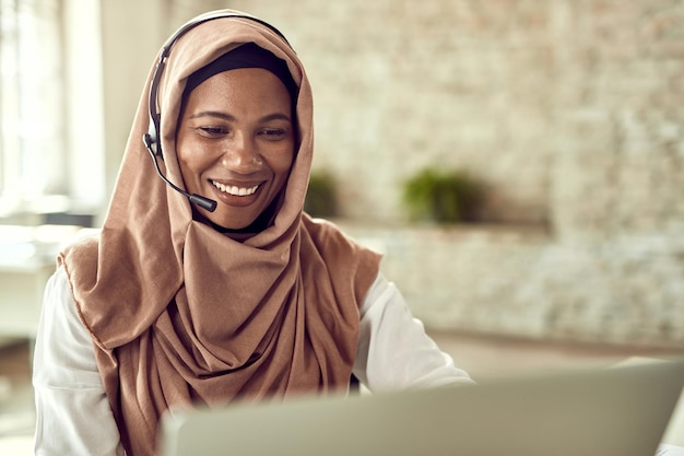 Feliz representante de servicio al cliente islámico hablando con su cliente mientras trabaja en una computadora en la oficina