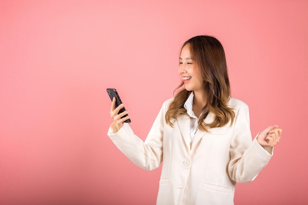 Feliz retrato asiático bonito jovem adolescente sorridente rosto animado usando telefone celular diga sim! foto de estúdio isolada em fundo rosa, mulher tailandesa surpresa faz gesto vencedor no smartphone