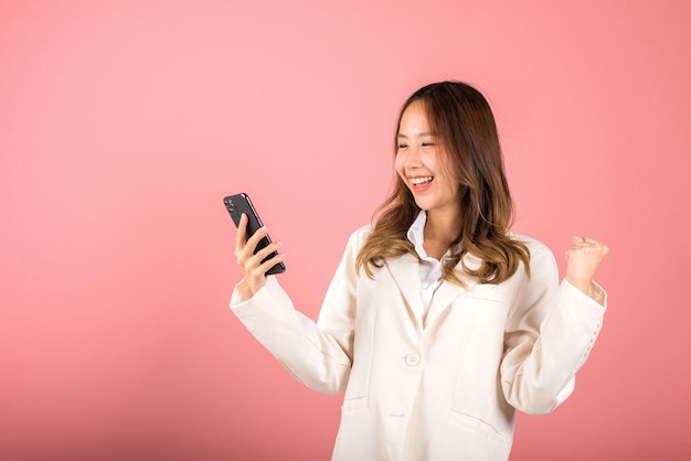 Feliz retrato asiático bonito jovem adolescente sorridente rosto animado usando telefone celular diga sim! Foto de estúdio isolada em fundo rosa, mulher tailandesa surpresa faz gesto vencedor no smartphone