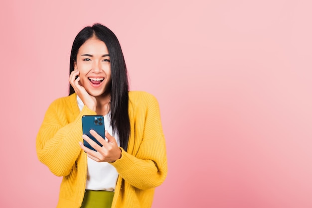 Feliz retrato asiático hermosa linda joven adolescente sonriendo emocionada usando un estudio de teléfono móvil inteligente aislado en fondo rosa Mujer tailandesa sorprendida haciendo gesto de ganador en el teléfono inteligente