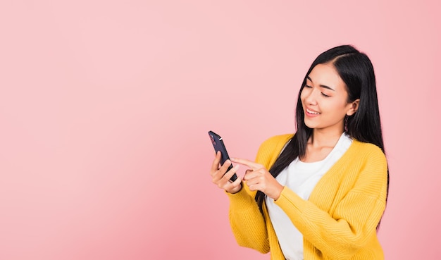 Feliz retrato asiático linda jovem adolescente sorridente animada digitando mensagem de texto no celular inteligente desfruta de comunicação on-line isolada, tiro em estúdio em fundo rosa com espaço de cópia
