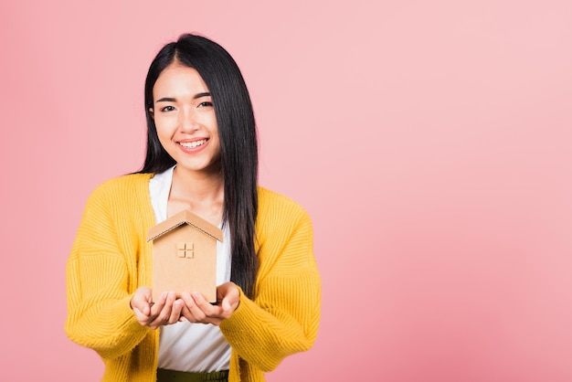 Feliz retrato asiático linda jovem bonita animada sorrindo segurando modelo de casa na mão, tiro de estúdio isolado em fundo rosa, corretora feminina segura seguro imobiliário em casa e conceito bancário
