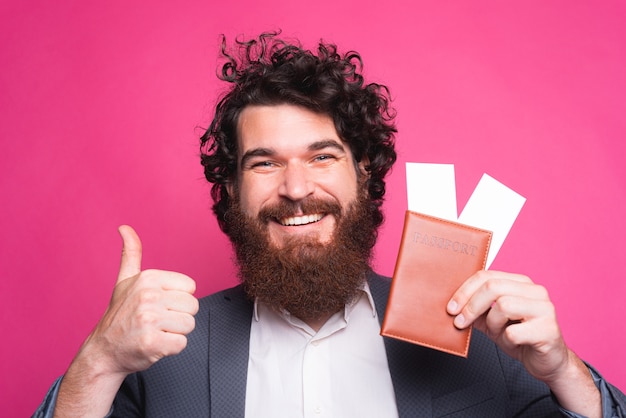 Feliz retrato de homem barbudo bonito em casual mostrando o polegar e passagens com passaporte