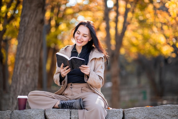 Feliz retrato de uma mulher estilosa no outono com sobretudo ela está bebendo café e lendo livro