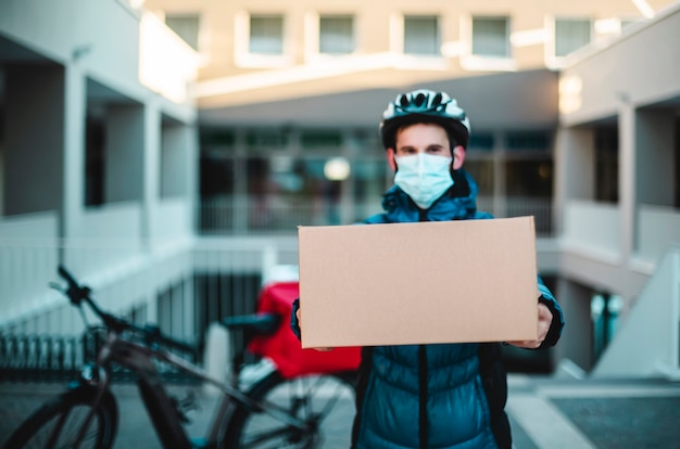 Feliz retrato do mensageiro com o pacote a ser entregue. O homem guarda a máscara protetora para o trabalho. Fundo de bicicleta. Entrega em domicílio, transportador, vírus