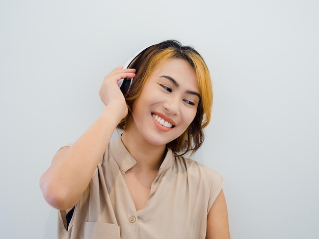 Feliz retrato de mujer asiática con cabello corto en camisa beige sin mangas disfruta escuchando podcast de música o canción en la radio con auriculares blancos sobre fondo blanco