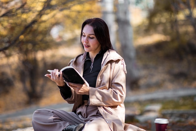 Feliz retrato de mujer con estilo en la temporada de otoño con gabardina, está tomando café y leyendo un libro