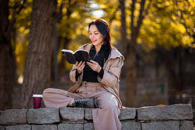 Feliz retrato de mujer con estilo en la temporada de otoño con gabardina, está tomando café y leyendo un libro