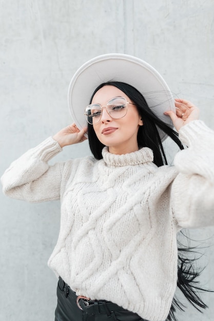 Feliz retrato urbano de una chica de moda con gafas antiguas en un suéter de punto de moda que se pone un sombrero en la calle cerca de una pared de hormigón gris