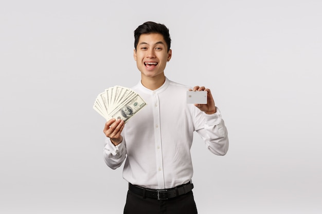 Feliz y rico, exitoso chico asiático en traje formal, con efectivo y tarjeta de crédito, riendo y sonriendo, alardeando de estabilidad financiera, tiene dos variantes de pago, banco de selección