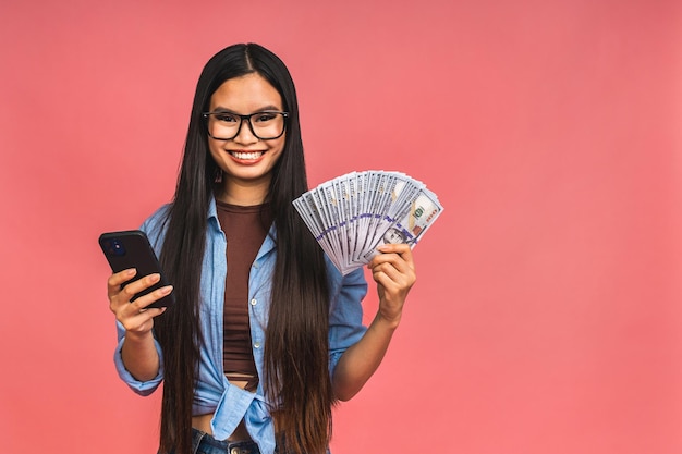 Feliz rico ganador Retrato de una mujer asiática sosteniendo billetes de dólares y sonriendo satisfecho de pie aislado sobre fondo rosa Usando teléfono móvil