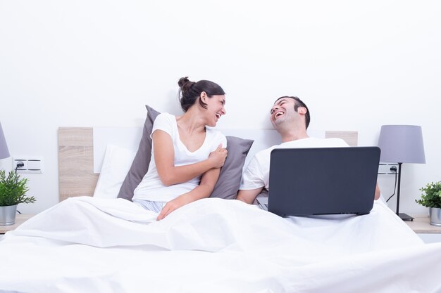 Feliz riendo relajado joven pareja usando la computadora portátil en la cama en su casa