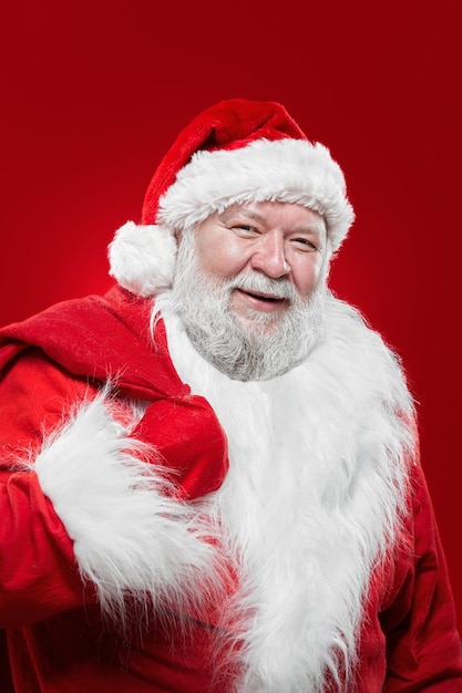 Feliz riendo Santa Claus con un abrigo rojo y un sombrero, mantenga una bolsa en la espalda, mirando a la cámara, fondo rojo aislado.