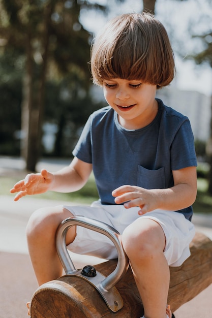 Feliz rindo menino em uma camiseta azul e shorts brancos passeios em uma cadeira de balanço de madeira ao ar livre. criança está brincando em um playground público em um dia ensolarado de verão. estilo de vida. espaço para texto