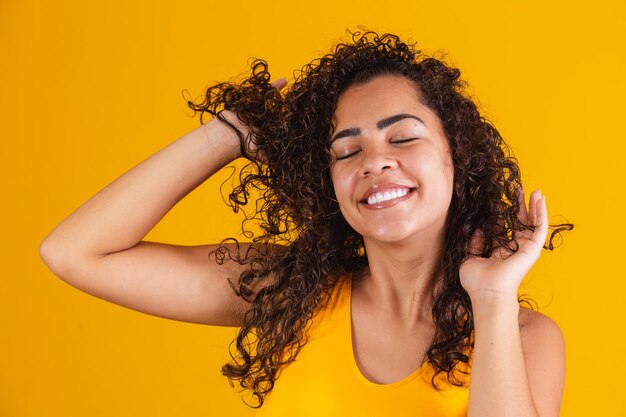 Feliz risa mujer afroamericana con su pelo rizado sobre fondo amarillo. Riendo a una mujer rizada vestida de amarillo tocándose el pelo y mirando a la cámara.