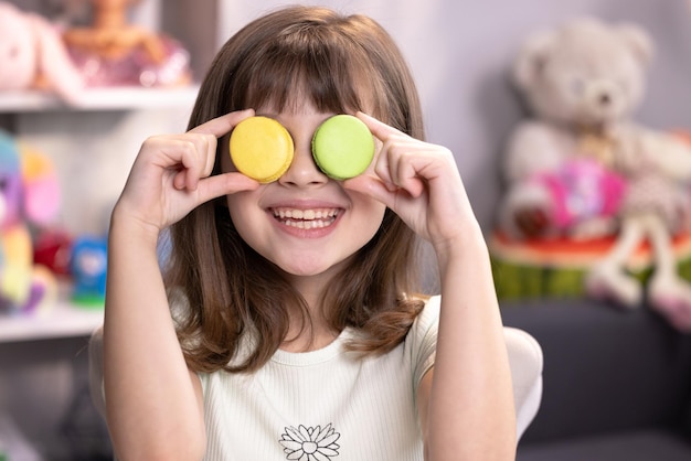 Feliz rosto sorridente de uma garotinha cobrindo os olhos com macaroons em um fundo colorido em casa