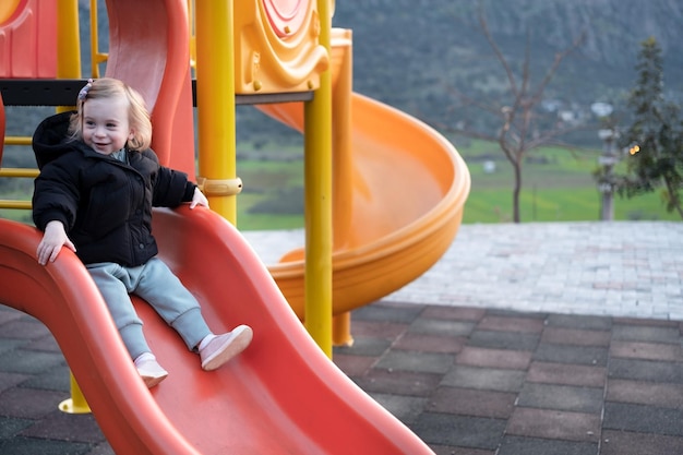 Feliz rubia caucásica niña bonita niño pequeño con ropa de abrigo deslizándose en el patio de recreo divirtiéndose Juegos infantiles para niños tiempo libre al aire libre estilo de vida Espacio de copia