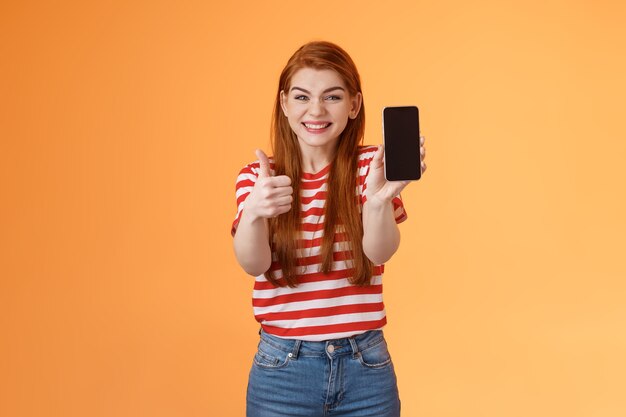 Foto feliz satisfecha pelirroja atractiva chica animando mostrar thumbup recomendar aplicación de teléfono inteligente mostrar teléfono móvil s ...