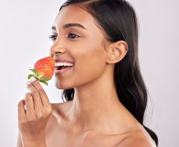 Foto feliz saudável e uma mulher comendo um morango para nutrição isolado em um fundo branco em um estúdio sorria pensando e uma garota indiana com uma fruta para um café da manhã dietético ou vitaminas de alimentos