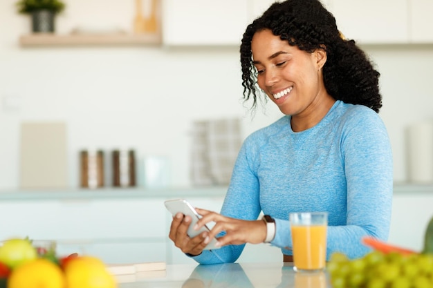 Feliz senhora afro-americana em roupas esportivas usando smartphone moderno enquanto bebe suco sentado na cozinha